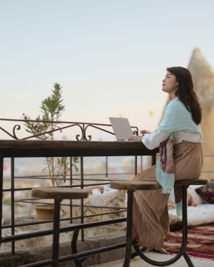 A photo of a woman sitting on a balcony searching through her laptop. 