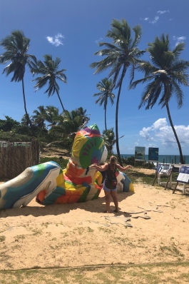 Alyssa Patton mimicking the posture of a very large and colorfully painted sea turtle sculpture on the beach