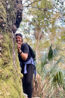 Alyssa Patton peeking around the edge of a large, lichen-covered tree trunk in the woods, with palmettos and scrubby trees in the background