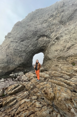 Amanda Pfeil standing at the base of a towering shoreside rock formation with a window in the rock directly behind her
