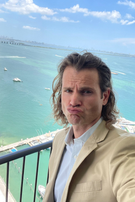 Andy Roberts on a balcony pursing his lips; the background shows a marina and the ocean in the distance