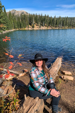 Angela Vaughn wearing a cow-person's hat sitting on a log in front of a wooded lake