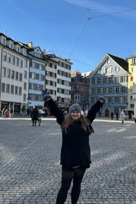 A photo of Angela with her hands up behind a Swiss city. 