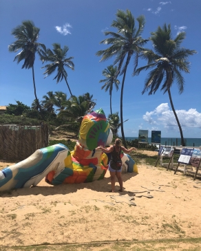 Alyssa Patton mimicking the posture of a very large and colorfully painted sea turtle sculpture on the beach