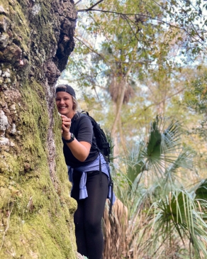 Alyssa Patton peeking around the edge of a large, lichen-covered tree trunk in the woods, with palmettos and scrubby trees in the background