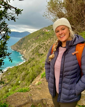 Alyssa Patton on a cliffside trail in Italy, with bright-bluish turquoise ocean in the background
