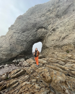 Amanda Pfeil standing at the base of a towering shoreside rock formation with a window in the rock directly behind her