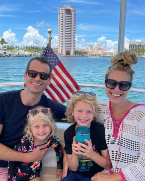 Andy Roberts and partner with two daughters on a boat with bright blue-green water and tall buildings behind them