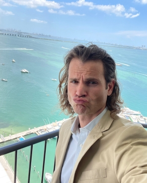 Andy Roberts on a balcony pursing his lips; the background shows a marina and the ocean in the distance