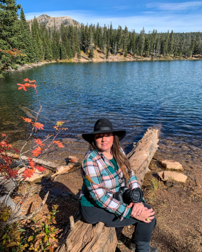 Angela Vaughn wearing a cow-person's hat sitting on a log in front of a wooded lake