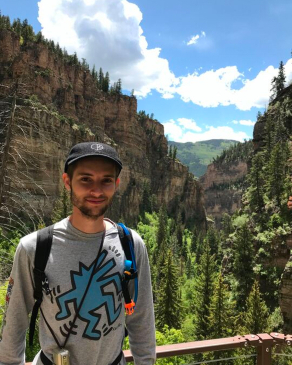 Ant Scalf on a cliffside lookout with rocky mountains and trees behind him