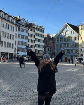 A photo of Angela with her hands up behind a Swiss city. 