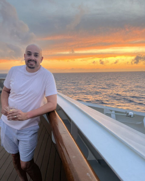 Mike smiles in front of a orange sunset while onboard a cruise. 
